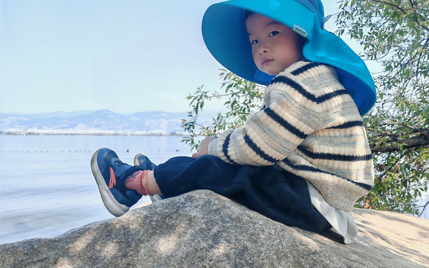 睿达戴着蓝色大帽子坐在湖边石头上